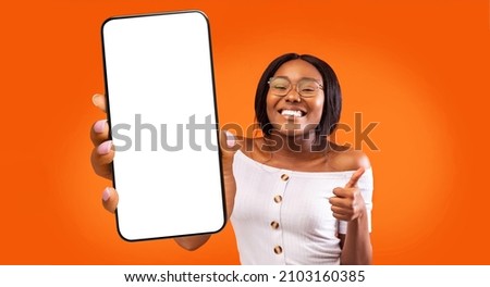 Great app. Overjoyed black lady holding big smartphone with white blank screen and showing thumb up, recommending mobile app or website, standing over orange background, mockup, panorama Royalty-Free Stock Photo #2103160385