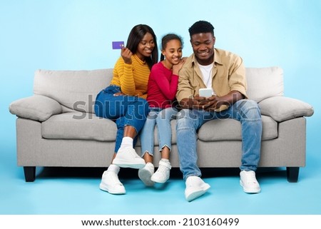 Online Shopping Concept. Happy Young Black Parents With Daughter Using Cell Phone And Debit Credit Card Buying Goods In Internet Web Store, Three People Sitting On Couch Isolated On Blue Studio Wall Royalty-Free Stock Photo #2103160499