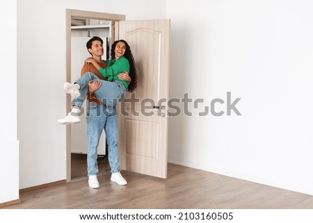 Mortgage And Relocation. Loving husband lifting carrying excited wife in his arms, holding lady on hands walking in new apartment, newlyweds celebrating moving day, having fun looking at empty room Royalty-Free Stock Photo #2103160505