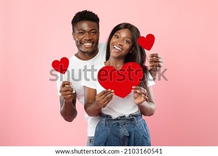 St. Valentines Day Concept. Portrait of African American couple in love holding red paper heart cards, smiling guy standing behind woman's back, looking at camera, isolated on pink studio background