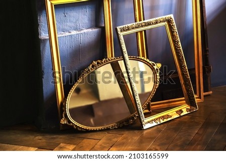 Gold picture frames and a retro oval mirror are arranged in a heap against a backdrop of wood floor and dirty wall. Close-up