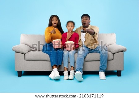 Emotional black family watching movie on TV and eating popcorn, excited spouses sitting on couch with daughter covering open mouth, surprised mom pointing at camera, isolated on blue studio background Royalty-Free Stock Photo #2103171119