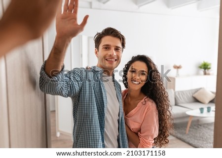 Portrait of cheerful couple welcoming inviting guests to enter home, happy young guy and lady standing hugging in doorway of modern flat, looking out together, man giving high five to visitor friend Royalty-Free Stock Photo #2103171158