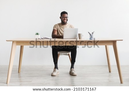 Happy young black man using laptop computer for online work at table in home office, free space. Cool African American guy having remote job, freelancing on web. Modern business concept Royalty-Free Stock Photo #2103171167