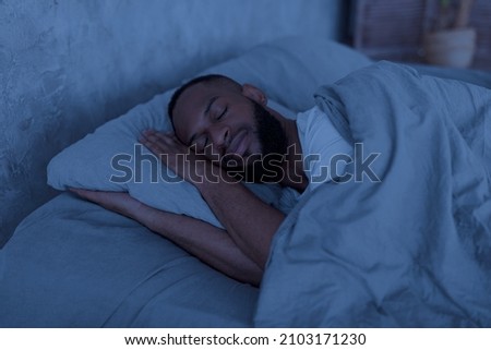 Healthy Sleeping Concept. Portrait of happy young well-slept African American man lying in bed with closed eyes, resting in bedroom on the side in the dark night, having hap