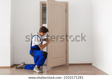 Maintenance Concept. Focused serious young locksmith with tattoo on hand wearing blue uniform standing on one knee in doorway installing doorknob handle, repairing wooden front door entrance Royalty-Free Stock Photo #2103171281
