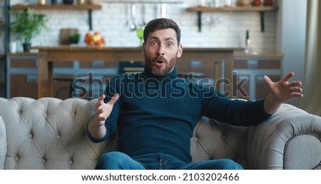 Amazed bearded man shocked, saying WOW, stunned by incredible news. Handsome male surprised to camera while sitting on sofa at home.  Royalty-Free Stock Photo #2103202466