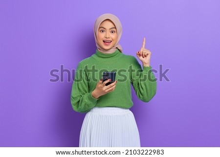 Excited young Asian woman in green sweater and hijab holding mobile phone and pointing finger up, creating genius solutions isolated over purple background Royalty-Free Stock Photo #2103222983
