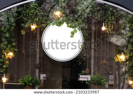 Blank minimal circular shop signboard mockup for design. Street hanging sign board for logo presentation with light garland amond the leaves. Metal cafe restaurant or bar badge black white round. Royalty-Free Stock Photo #2103233987
