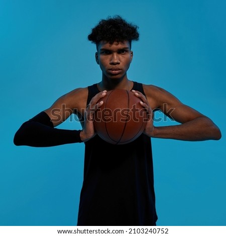 Black serious basketball player holding basketball ball and looking at camera. Concept of healthy lifestyle. Young curly athletic man wearing black tank top. Isolated on blue background. Studio shoot