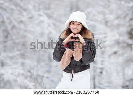 Portrait of Girl in Winter nature showing Valentine heart shape and smiling. Valentine's Day. Happy Teenage Model Girl having fun in snow Forest, enjoying and laughing outdoors. Love