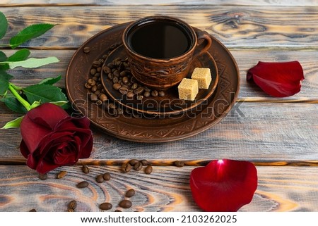 A clay cup with black coffee on a saucer on a wooden table. Red rose and coffee beans in the background. Rose petals on the table. Light wooden background