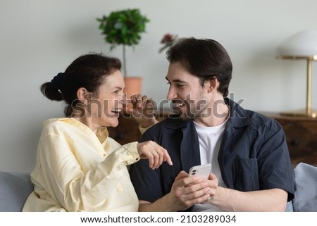 Happy young man teaching laughing mature senior mother using cellphone applications, watching funny photo video content online in social network, shopping in internet store, modern tech addiction. Royalty-Free Stock Photo #2103289034