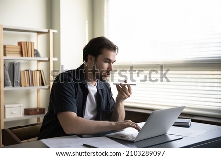 Focused young male employee entrepreneur recording audio message on cellphone or holding loudspeaker conversation, multitasking web surfing information working on computer in modern home office.