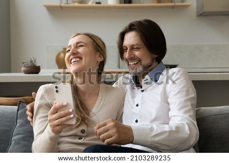 Cheerful excited 40s couple using mobile phone at home, laughing out loud, having fun. Adult husband and wife sharing device, shopping on internet, chatting online, getting good funny news