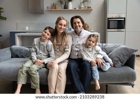 Happy parents and two sibling children relaxing, hugging on sofa at home, looking at camera, smiling. Millennial family couple and kids posing on comfortable couch in new apartment front portrait