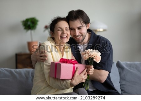 Joyful bonding middle aged senior woman feeling excited getting wrapped gift box and flowers from young smiling grownup son, sitting together on cozy couch, birthday or special occasion celebration.