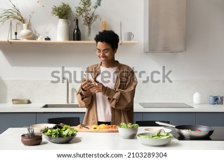 Happy Black food blogger girl taking picture of salad ingredients on mobile phones, recording video, shooting cooking process, reading recipe on Internet, chatting online in home kitchen Royalty-Free Stock Photo #2103289304
