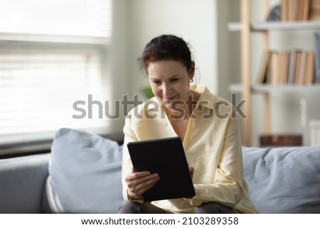 Happy beautiful old mature retired woman using digital touchpad gadget, communicating in social network, shopping online, web surfing information or playing games, sitting on comfortable sofa.