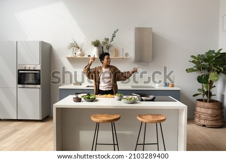 Happy joyful African dancer girl listening to music, dancing while preparing dinner in home kitchen, having fun at cooking island table with cut vegetables, healthy organic food ingredients Royalty-Free Stock Photo #2103289400