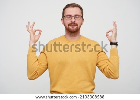 Studio portrait of young bearded male student wears yellow sweater keeps calm, keeps eyes closed and showing yoga mudra sign, isolated over white background