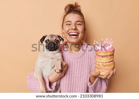 Positive birthday girl celebrates bday together with favorite dog has upbeat mood holds doughnuts with burning candles pug dog wears knitted sweater isolated over beige background. Holiday concept