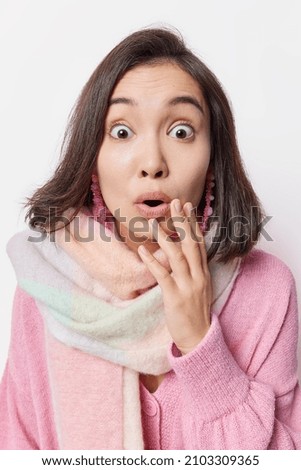 Surprised scared young Asian woman with dark hair stares bugged eyes holds breath keeps mouth opened cannot believe in shocking news wears jumper and scarf around neck poses indoor. Omg concept