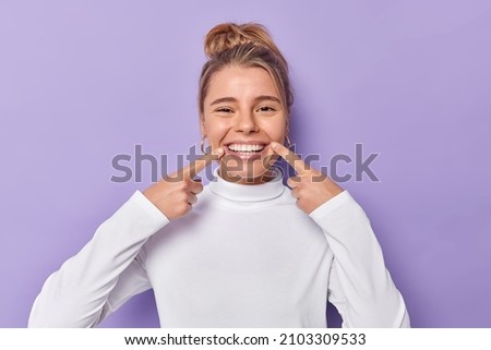 Dental care concept. Happy young fair haired woman points index fingers at toothy smile shows wel cared white teeth after whitening or dentist visit dressed casually isolated over purple background Royalty-Free Stock Photo #2103309533