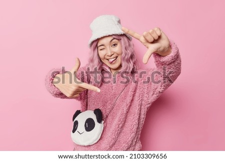 Positive carefree Asian woman winks eye sticks out tongue makes frame gesture finds perfect angle wears white fur panama and coat isolated over pink background gazes at camera through hands.