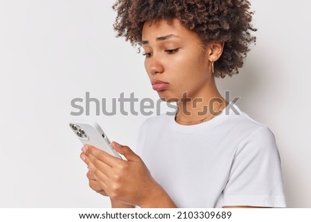 Upset curly haired woman feels sad and unhappy reads sms on smartphone grimaces with frustraated expression receives disappointing news via message complains about something wears casual t shirt