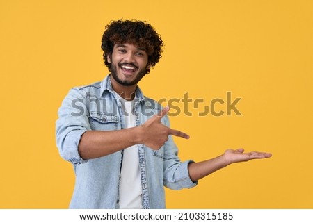 Excited happy positive young indian man student pointing aside with fingers hand gesture at copy space advertising product, presenting sale discount promo offer standing isolated on yellow background. Royalty-Free Stock Photo #2103315185