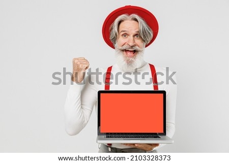Elderly gray-haired mustache bearded man 50s in turtleneck red hat suspenders hold use work laptop pc computer blank screen workspace area do winner gesture isolated on plain white background studio