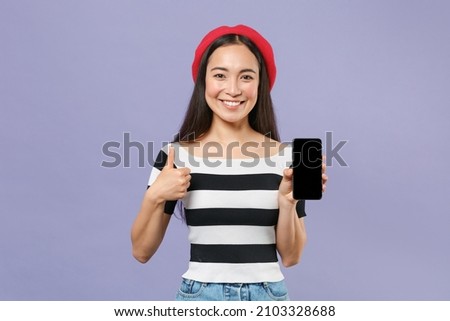 Smiling young asian woman wearing striped t-shirt red beret hold mobile cell phone with blank empty screen mock up copy space showing thumb up isolated on pastel violet background studio portrait