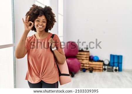 African american woman with afro hair holding yoga mat at pilates room smiling positive doing ok sign with hand and fingers. successful expression. 