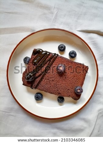 hdr, It's a picture of cake slice with blueberries and chocolate syrupy, on a white plate and white background. 