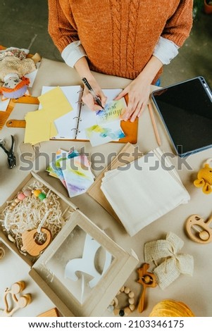 Female crafter writing a card with gratitude for the buyer. Young woman packing purchases at eco friendly package at her workshop of handmade kids toys. Top view, flat lay.