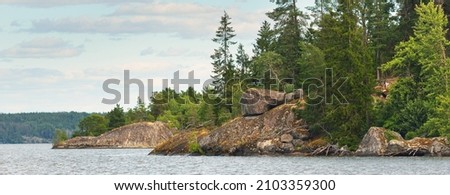 Rocky lakeshore and forest on a sunny summer day. Green deciduous trees, plants. Strängnäs, Mälaren lake, Sweden. Idyllic landscape. Nature, ecology, environment, ecotourism Royalty-Free Stock Photo #2103359300