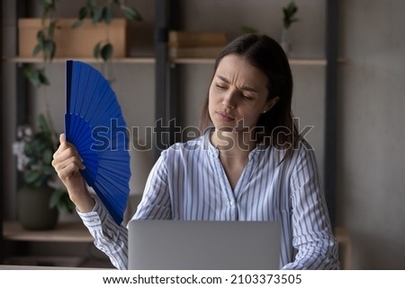 Exhausted office employee suffering from heat, hot stuffy air, working at laptop from home without conditioner on summer day. Student girl, business woman cooling with blue handheld fan at workplace Royalty-Free Stock Photo #2103373505