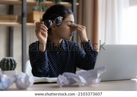 Tired millennial Indian business woman feeling headache, suffering from dry irritated eyes due to overload with paperwork,. Sad office employee touching head, eyelids at laptop, thrown crumpled papers Royalty-Free Stock Photo #2103373607