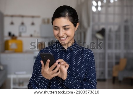 Happy millennial Indian girl, young woman reading text message on smartphone screen, smiling, feeling joy, chatting, typing, using online service, virtual app for payment on internet at home Royalty-Free Stock Photo #2103373610