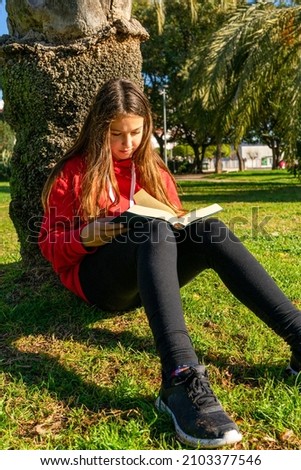Pretty Hispanic girl sitting on the grass in a park reading a book on a sunny winter day. Leisure concept