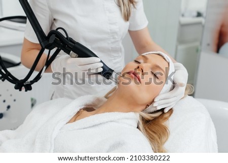 A young girl gets a charcoal peeling for the skin of the face in a beauty salon. Laser pulses cleanse the skin. Hardware cosmetology. The process of photothermolysis, warming the skin. Royalty-Free Stock Photo #2103380222