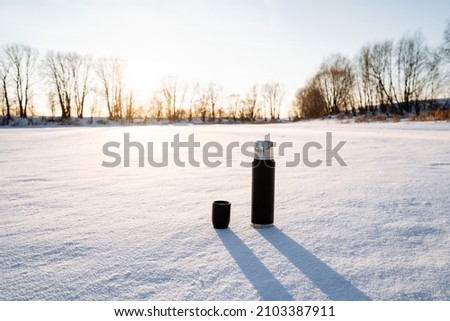 A thermos of black with a cup stands on the snow. Tourist utensils. Drink hot tea in winter to keep warm. Vacuum bottle for hot water. Glare of the sun in nature. High quality photo