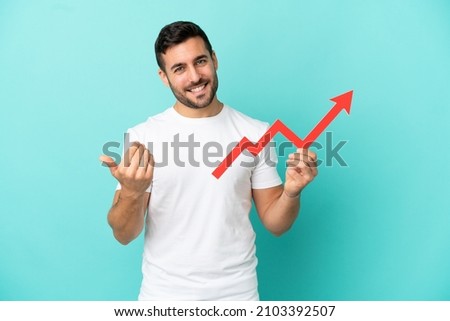 Young handsome caucasian man isolated on blue background holding a catching a rising arrow and doing coming gesture Royalty-Free Stock Photo #2103392507