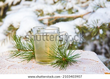 Hot pine tree needle tea infusion in transparent glass tea cup on table. Snowy pine tree on background, outdoors on cold winter day. Royalty-Free Stock Photo #2103403907