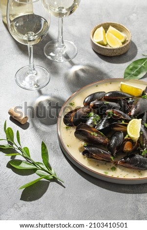 Black mussels in garlic sauce with herbs and lime and two glasses of white wine on the table under the summer sun, vertical photo Royalty-Free Stock Photo #2103410501