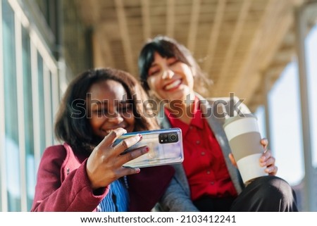 Two multiracial office worker women using a mobile phone to take a picture and having coffee on a break from work