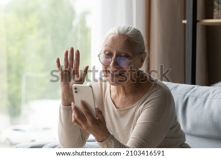 Happy old mature retired grandmother in eyeglasses waving hand, looking at smartphone web camera, starting video call distant conversation with grownup children or friends, chatting online at home. Royalty-Free Stock Photo #2103416591