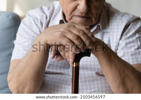 Close up cropped image frustrated unhappy elderly mature retired man with walking disability holding folded hands on wooden cane, thinking of physical health problems, retirement illnesses, arthritis. Royalty-Free Stock Photo #2103416597