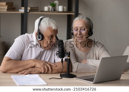 Happy middle aged old 70s family couple in headphones speaking in professional stand microphone, recording podcast or educational lecture online on computer, vlogging or communicating distantly.